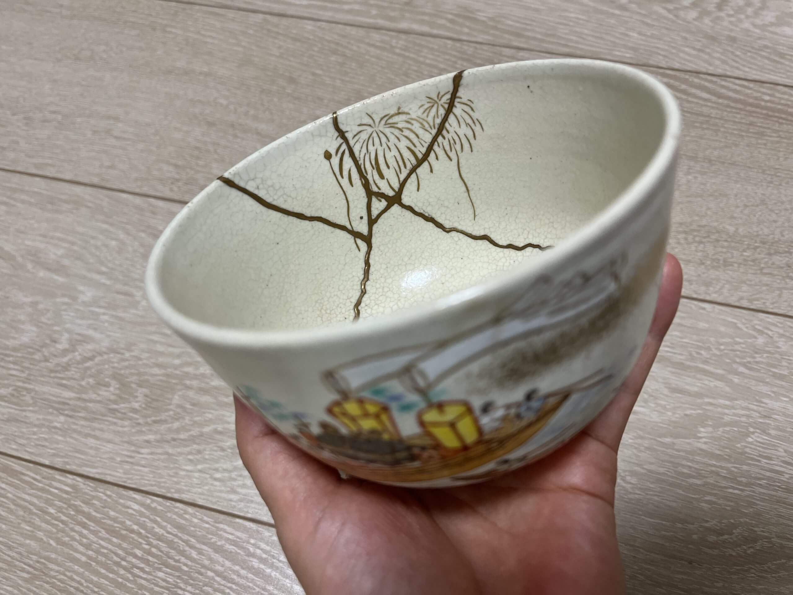 Japanese tea bowl repaired with kintsugi, showing gold-filled cracks and a colorful illustration of the Tenjin Matsuri, an annual festival in Osaka, on the front